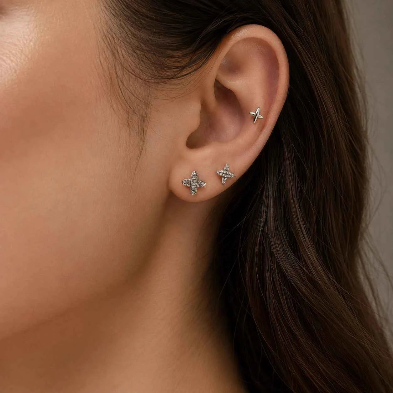 Silver-tone X-shaped and charm stud earrings on ear, side view