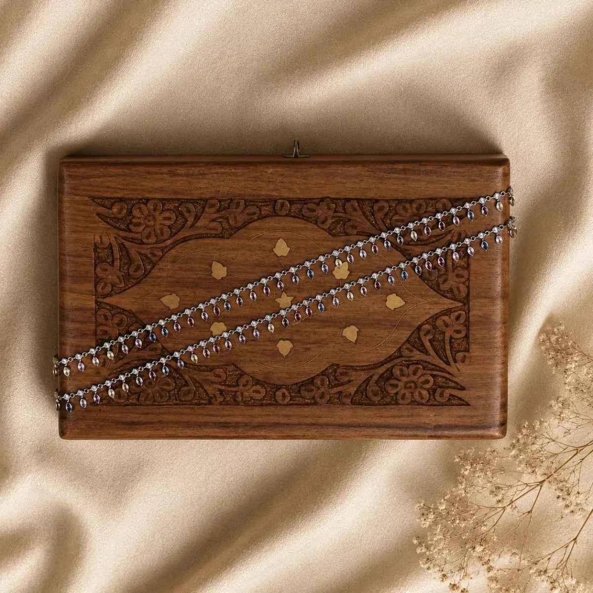 Silver chain anklet laid across a wooden box with decorative carvings